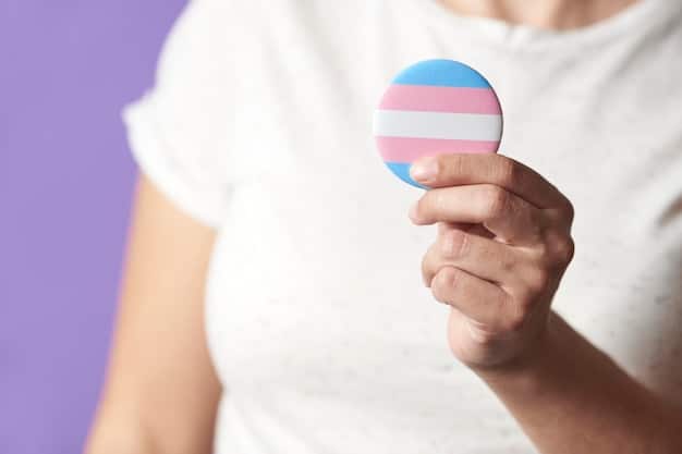 A close-up shot of a hand holding a transgender pride flag pin, with a gaming mouse visible in the blurred background. The focus is on the pin, symbolizing identity and representation within the gaming community.